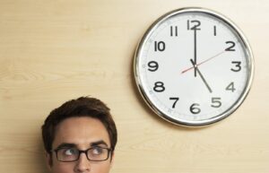 Young businessman wearing spectacles looking at clock on wooden wall in office